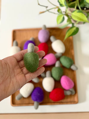 Hand holding single green felt lightbulb with tray of colorful felt lightbulbs on wooden tray below and plant nearby