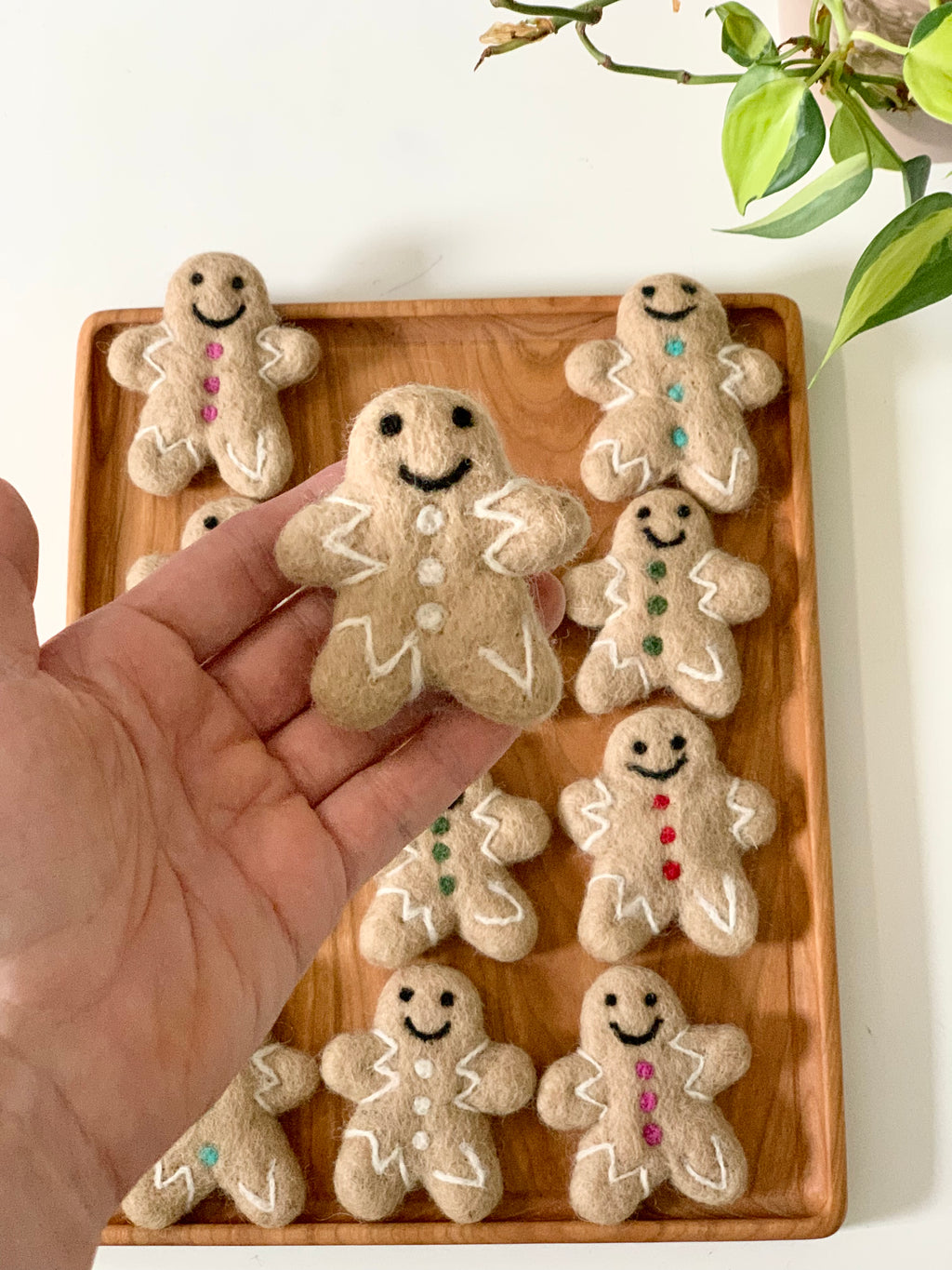 Handmade felt gingerbread person with stitched smile and buttons held above wooden tray with similar felt figures on white table