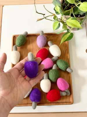 Hand holding a purple felt lightbulb with other colorful felt lightbulbs on wooden tray beside green plant on white table