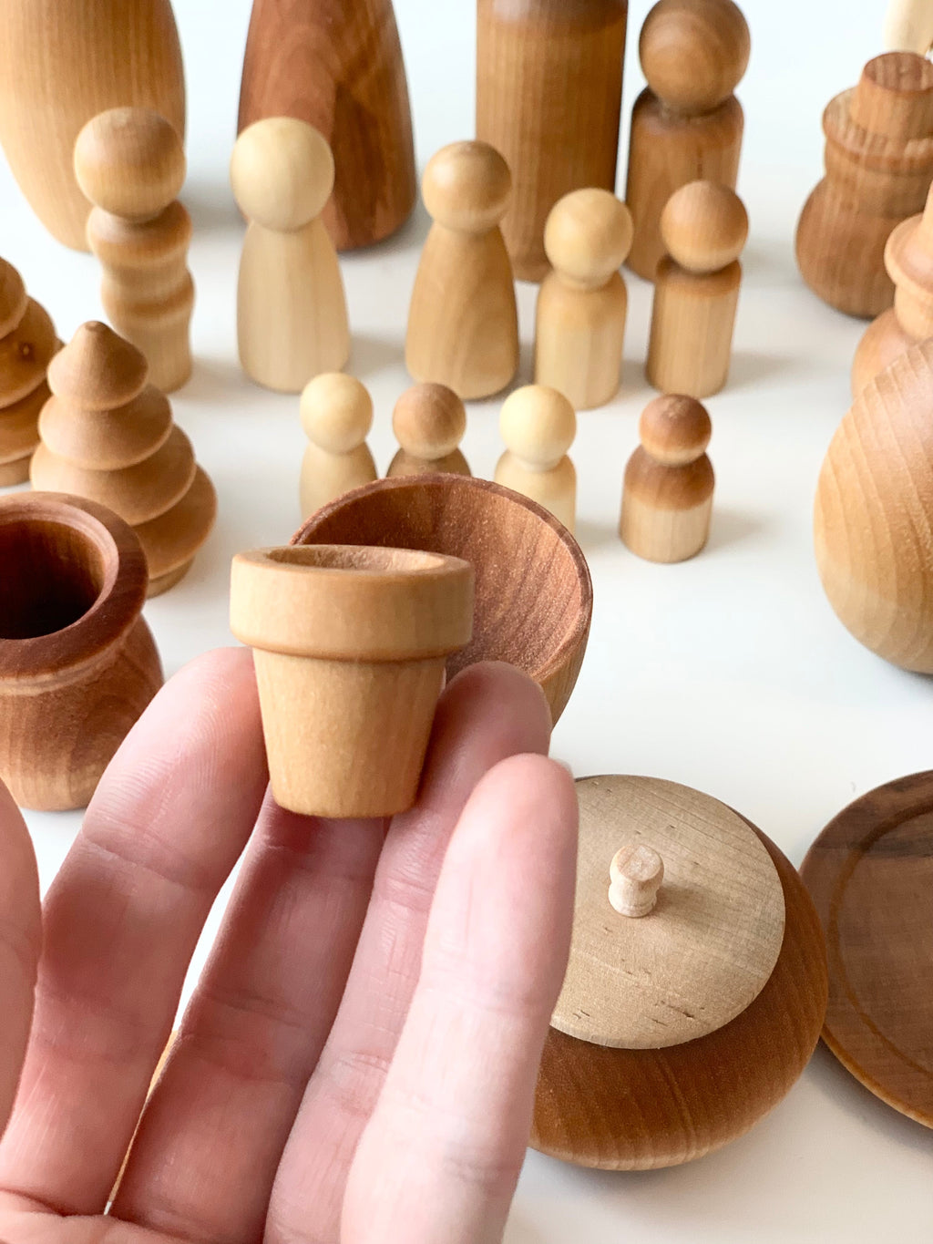 Hand holding small natural wooden pot with various smooth unfinished wooden figures and shapes on white surface