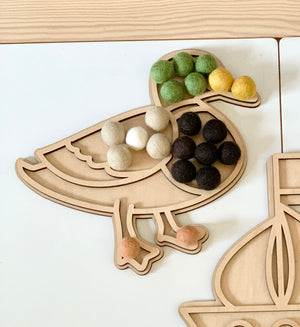 Maple plywood duck-shaped fillable play tray with green, yellow, beige, black, white, and peach felt balls on white table