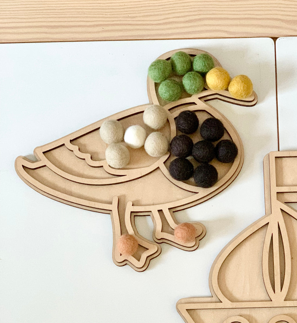 Maple plywood duck-shaped fillable play tray with green, yellow, beige, black, white, and peach felt balls on white table