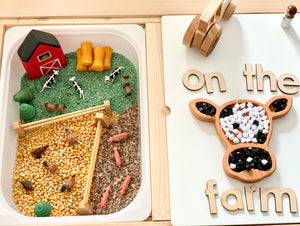 Wooden cow-shaped sensory tray with black, white, and wooden beads next to farm-themed play area with miniature animals and barn