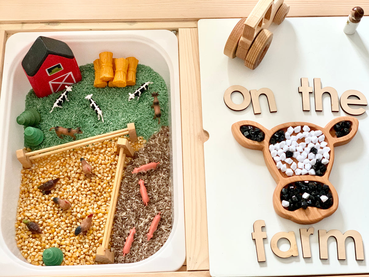 Wooden cow-shaped sensory tray with black, white, and wooden beads next to farm-themed play area with miniature animals and barn