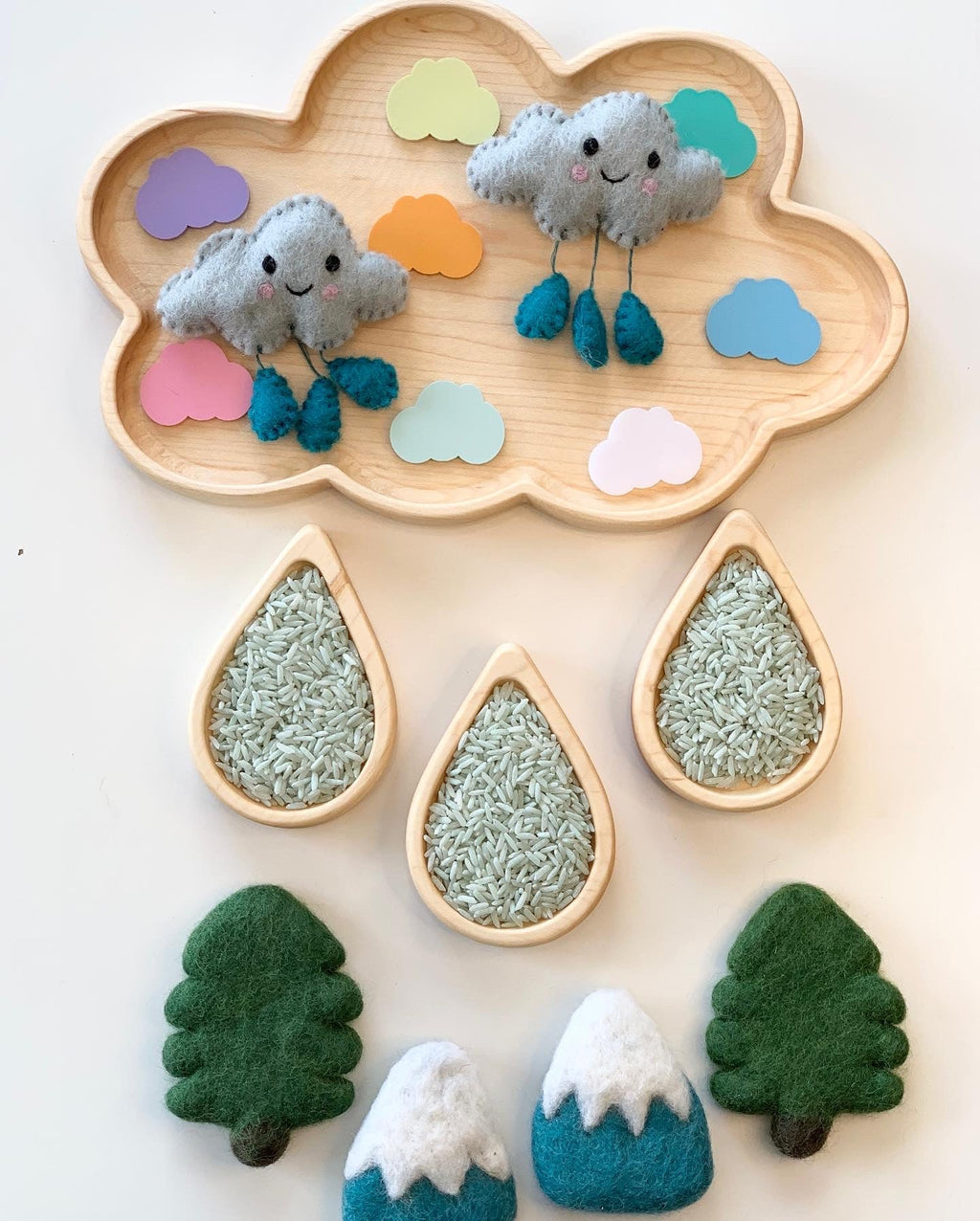 Wooden cloud-shaped tray with felt clouds, colorful paper clouds, wooden raindrop trays filled with rice, felt trees and snowy mountains arranged below.