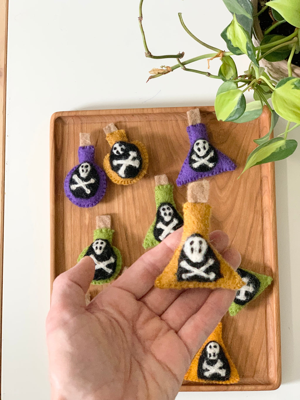 Handmade felt potion bottles in purple, orange, and green with skull and crossbones on wooden tray with hand holding one