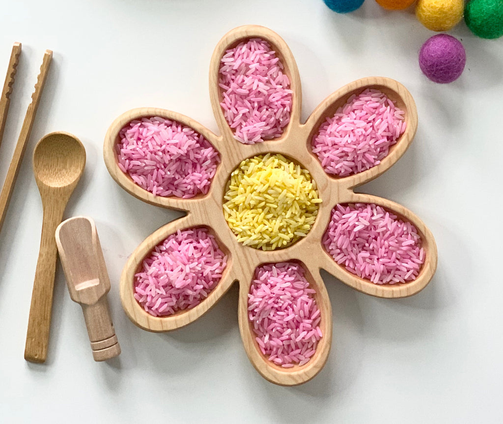 Wooden flower-shaped tray filled with pink and yellow colored rice next to wooden scoops and tongs on white surface