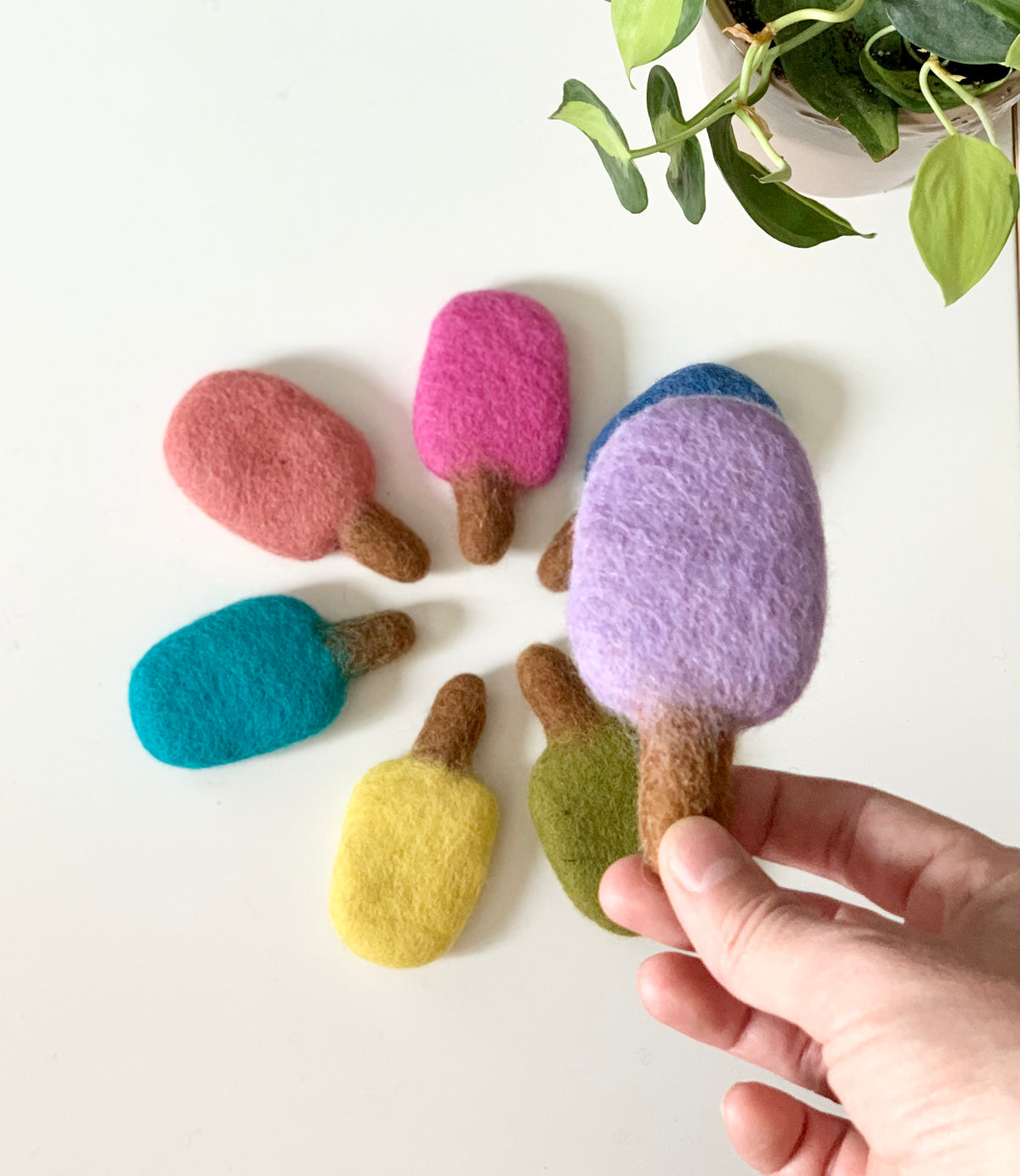 Hand holding a purple and blue felt popsicle with other colorful felt popsicles on white table with green plant nearby
