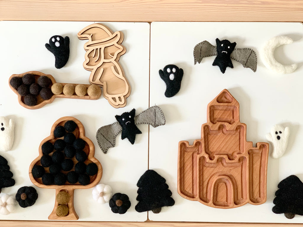 Wooden fillable witch play tray with felt ghosts, bats, and pom poms in witch, tree, broom, and castle shapes on white table