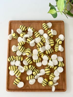 Multiple small yellow felt bees with black stripes and white wings scattered on a wooden tray next to a green plant