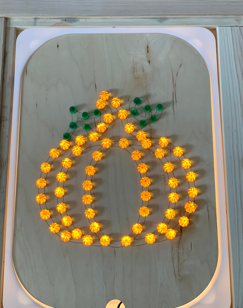 Wooden geometric board insert with warm orange lights shaped like a pumpkin with green leaf details on light wood surface