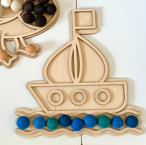 Wooden boat-shaped play tray with colorful blue and green felt balls arranged as waves on white surface