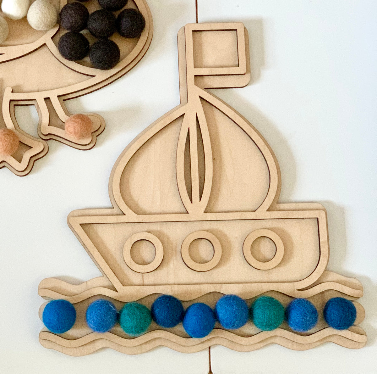 Wooden boat-shaped play tray with colorful blue and green felt balls arranged as waves on white surface