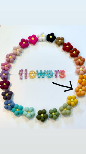Circle of colorful handmade felt flowers with yellow centers and glittery letters spelling flowers on white background