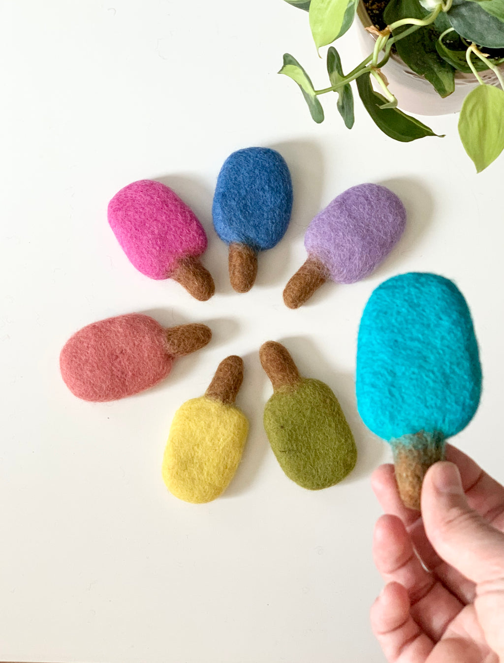 Handmade felt popsicles in various colors arranged in a circle on white surface with plant nearby