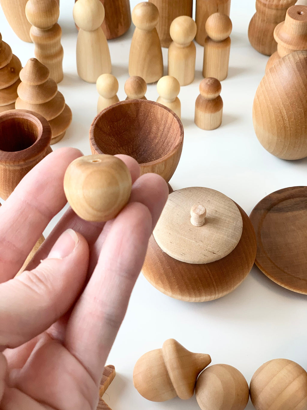 Hand holding small unfinished wooden apple shape with various smooth natural wood pieces and figures on white surface