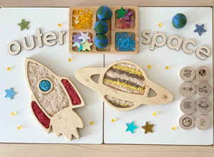 Wooden rocket and Saturn-shaped trays filled with colored sand surrounded by stars, planets, beads, and wooden letters spelling outer space