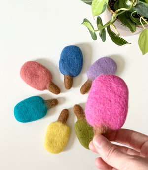 Handmade felt popsicles in pink, blue, purple, teal, yellow, green, and red on white surface with green plant