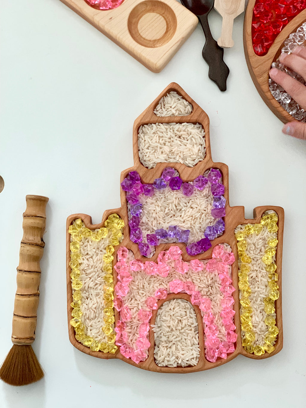 Castle-shaped wooden sensory tray filled with rice and colorful translucent beads on white table with brush nearby