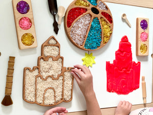 Castle-shaped wooden sensory tray filled with rice, colorful gem trays, and sandcastle mold on white table