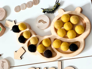 Wooden caterpillar-shaped sensory tray filled with yellow and black felt balls and a small caterpillar figure on white table
