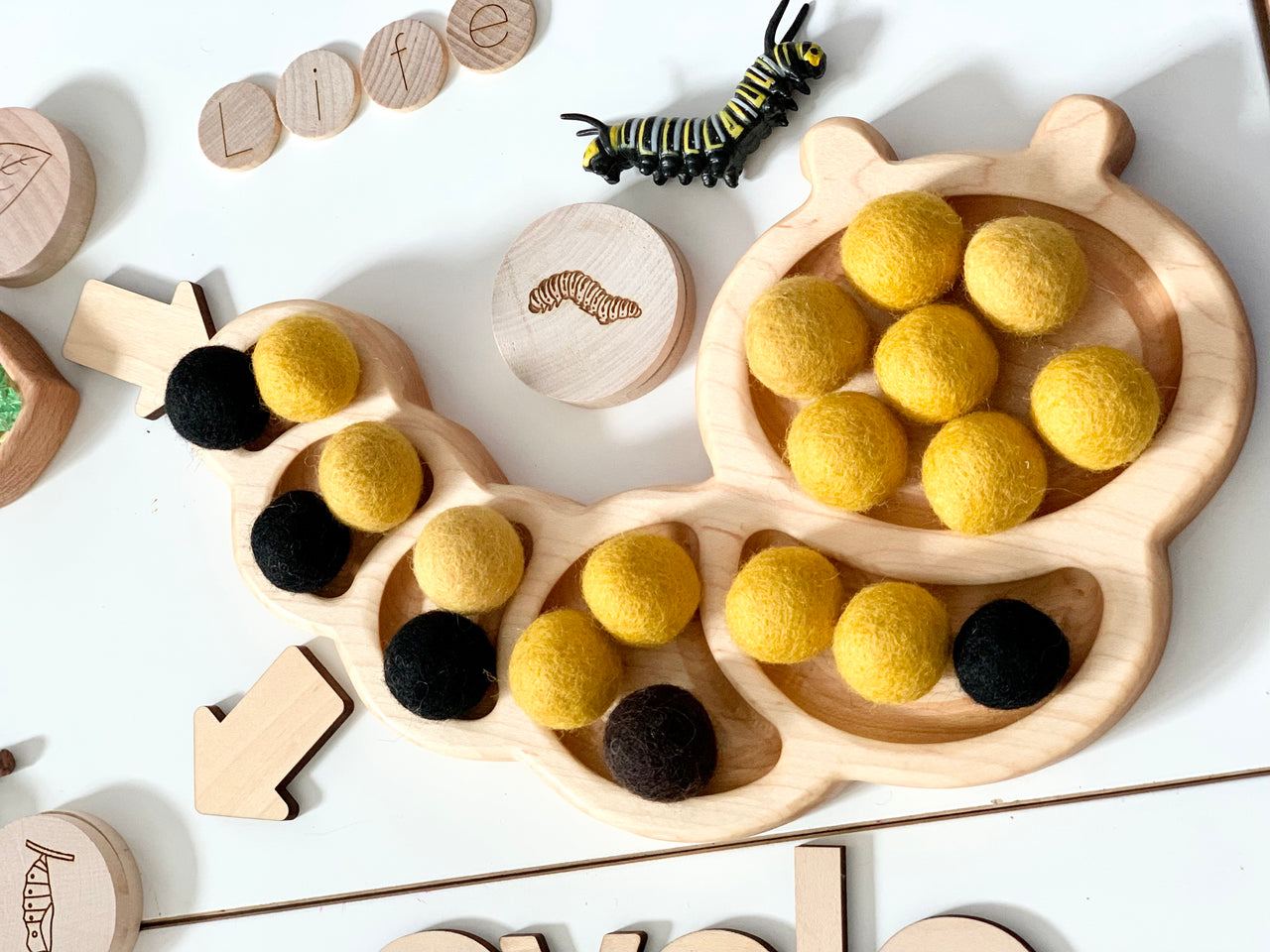 Wooden caterpillar-shaped sensory tray filled with yellow and black felt balls and a small caterpillar figure on white table