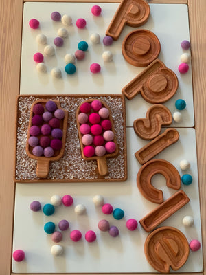 Wooden popsicle-shaped sensory tray with colorful felt balls and letters spelling popsicle on white and wooden surface