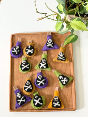 Handmade felt potion bottles in purple, orange, and green with skull and crossbones designs on wooden tray with plant nearby