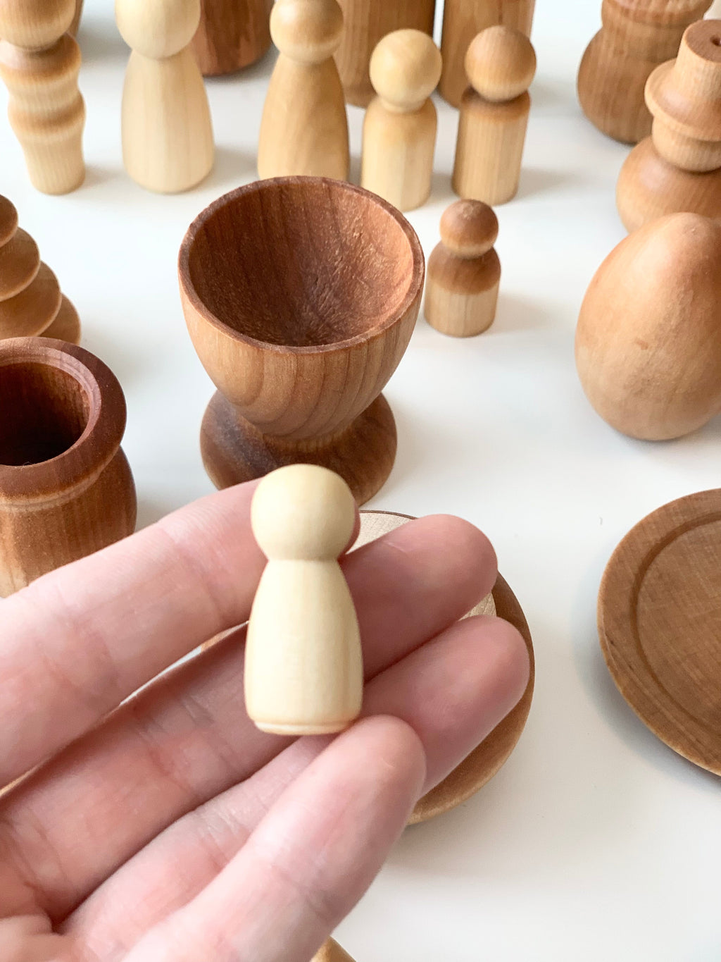 Hand holding small unfinished natural wood peg figure among various wooden cups and shapes on white surface