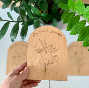 Hand holding personalized wooden garden plaque engraved with wildflowers and "Grannie's Garden" under green plant leaves