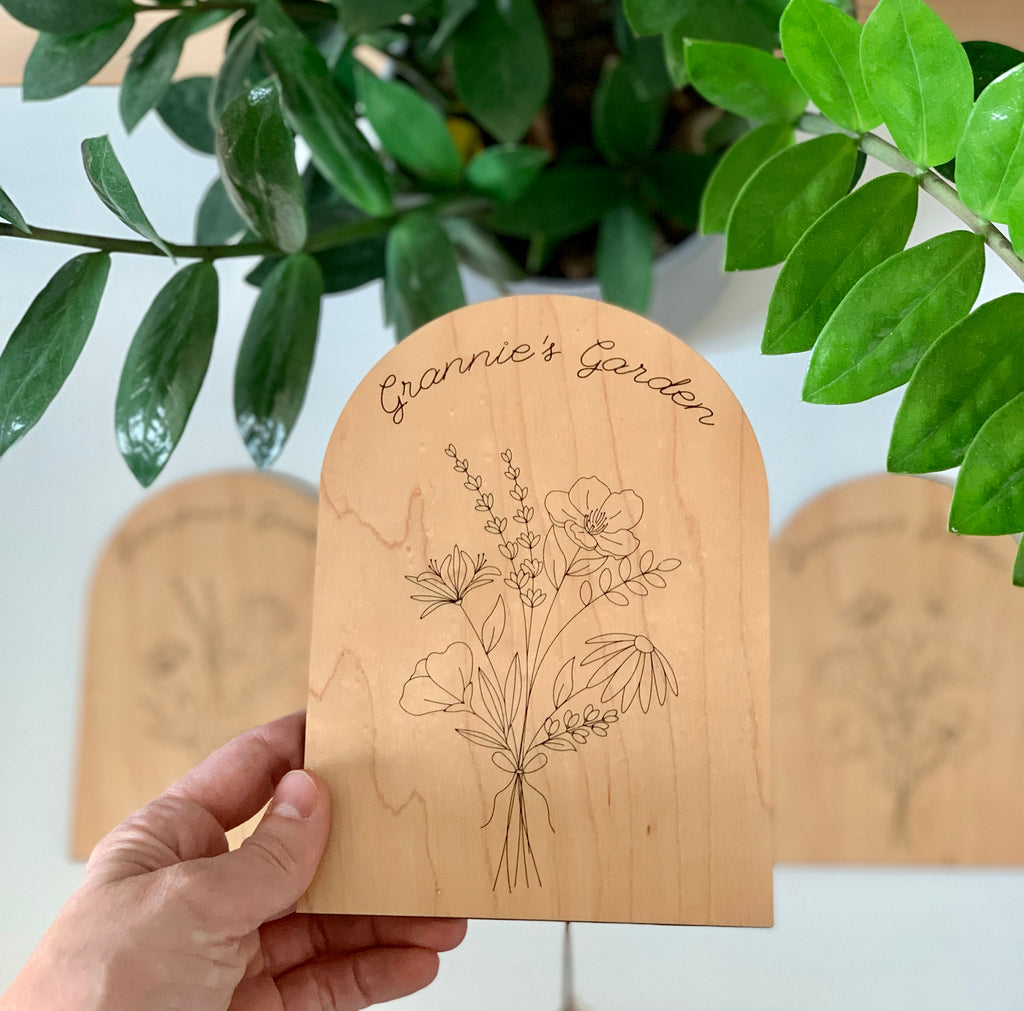Hand holding personalized wooden garden plaque engraved with wildflowers and "Grannie's Garden" under green plant leaves