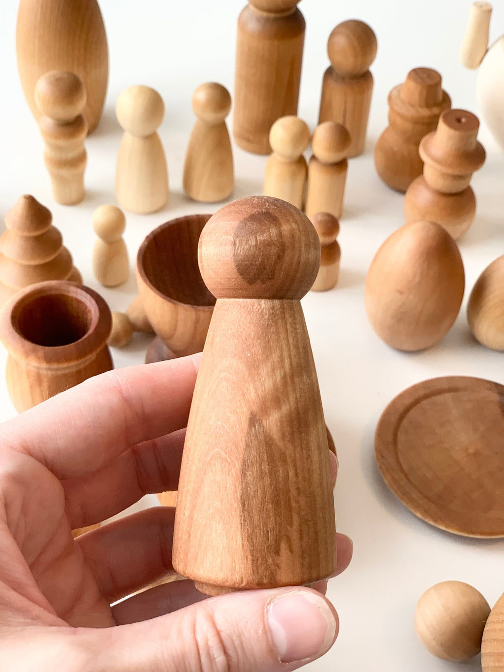 Hand holding tall natural wooden peg figure with various smooth unfinished wooden shapes and vessels on white surface