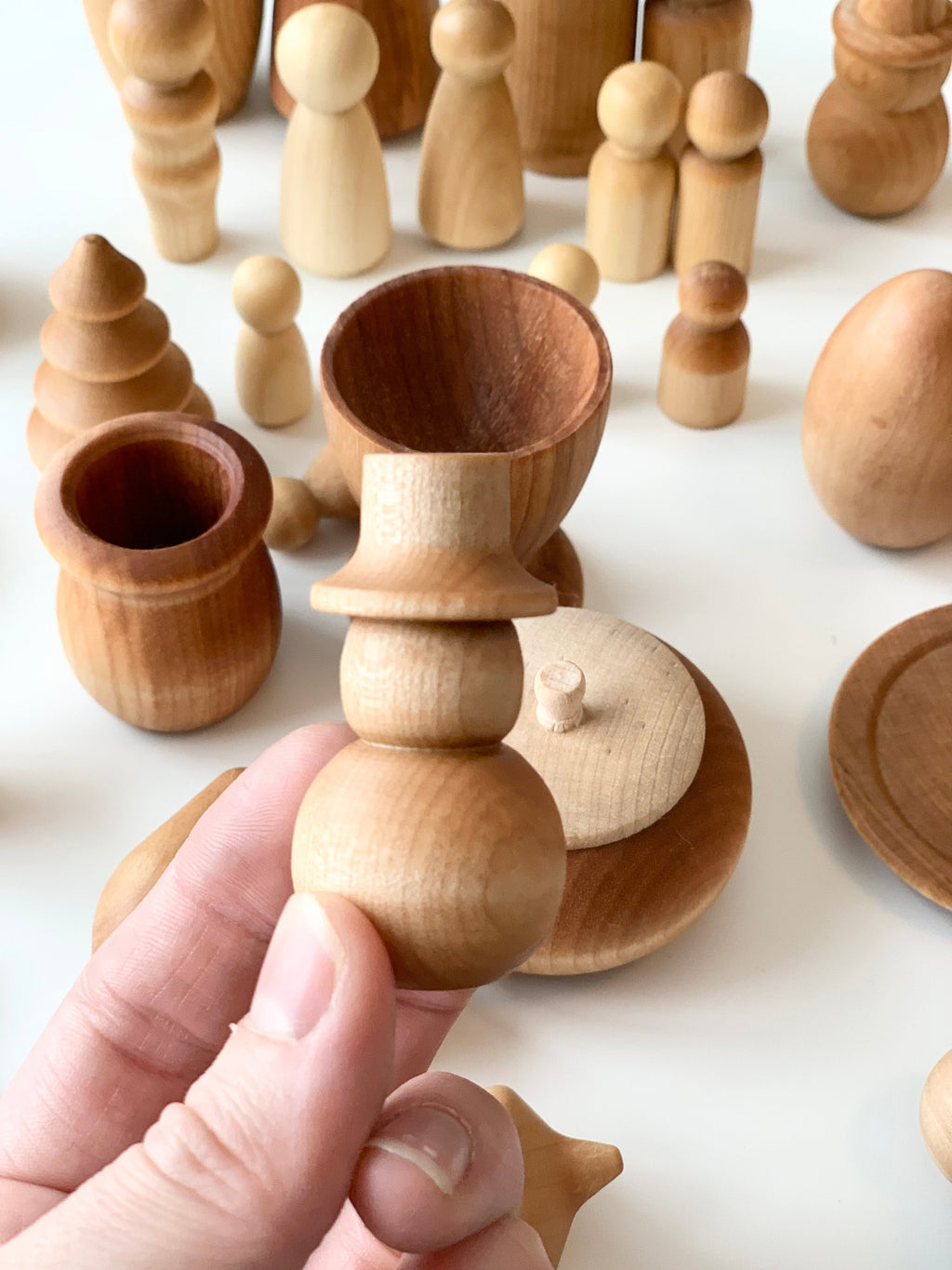 Hand holding a small natural wooden figurine with a top hat among various smooth wooden shapes and vessels on white surface