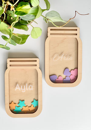 Two personalized wooden reward jars shaped like mason jars filled with pastel wooden tokens on white surface