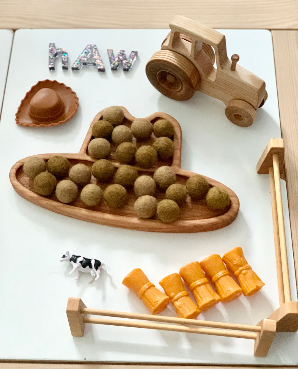 Wooden cowboy hat sensory tray filled with felt balls, miniature wooden tractor, small plastic cow, hay bales, and letter tiles spelling haw