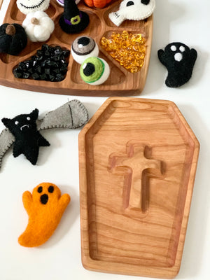 Wooden coffin-shaped sensory tray with cross cutout surrounded by felt Halloween figures on white surface