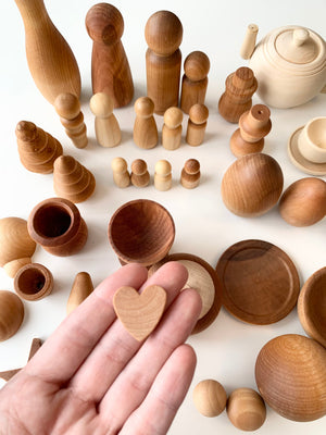 Hand holding small wooden heart shape with various unfinished wooden figures, bowls, and stacking toys on a white surface