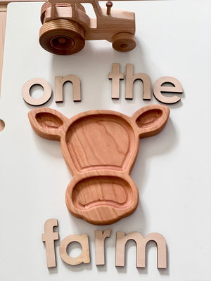 Wooden cow-shaped sensory tray with wooden letters spelling on the farm and a toy wooden tractor on white surface