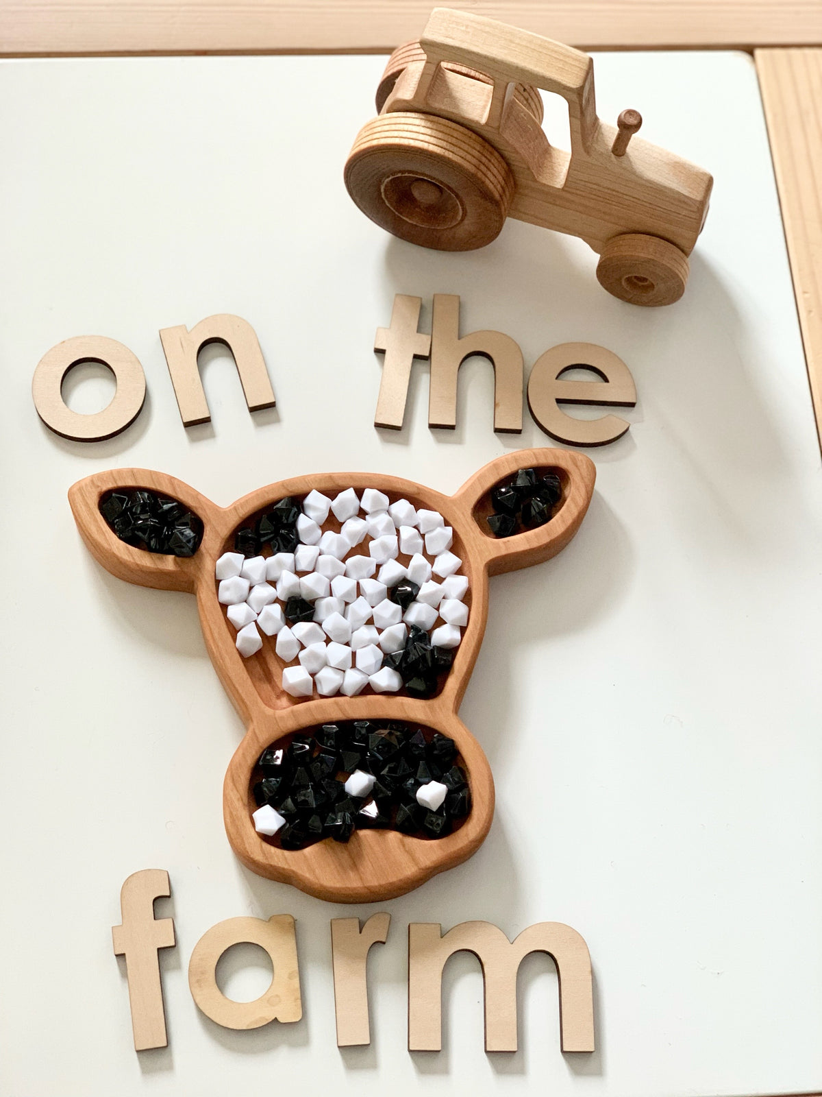Wooden cow-shaped sensory tray with black and white beads, wooden letters spelling on the farm, and wooden tractor toy on white surface