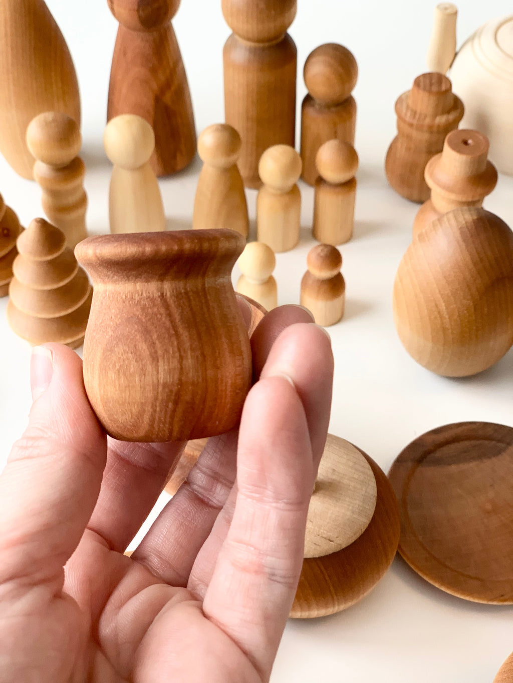 Hand holding small rounded smooth wooden figure with various unfinished wooden shapes and peg dolls in background