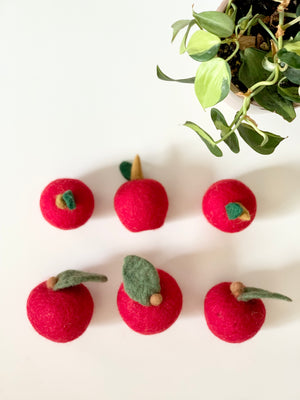 Six handmade red felt apples with small green leaves arranged on white surface next to green plant