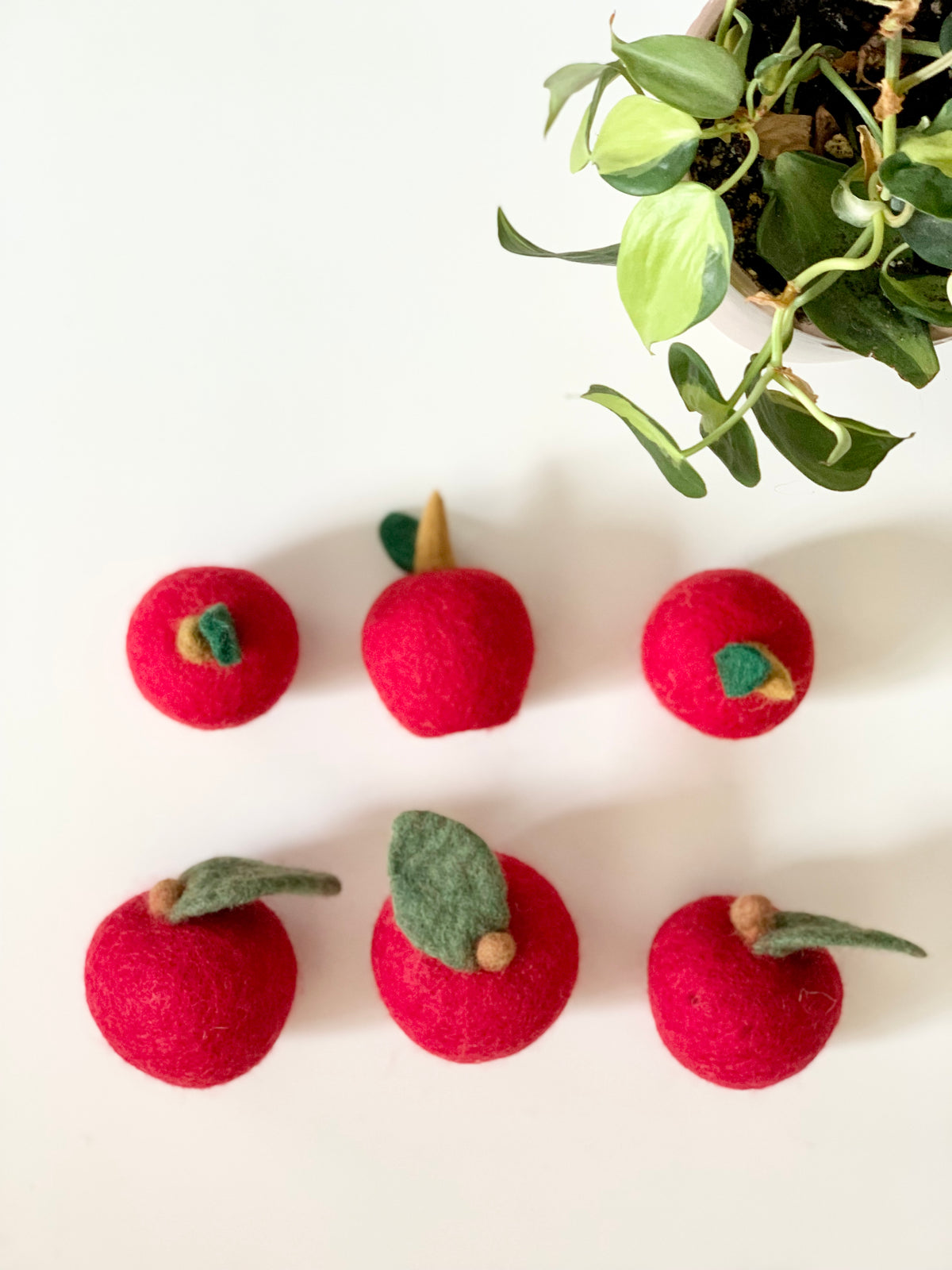 Six handmade red felt apples with small green leaves arranged on white surface next to green plant