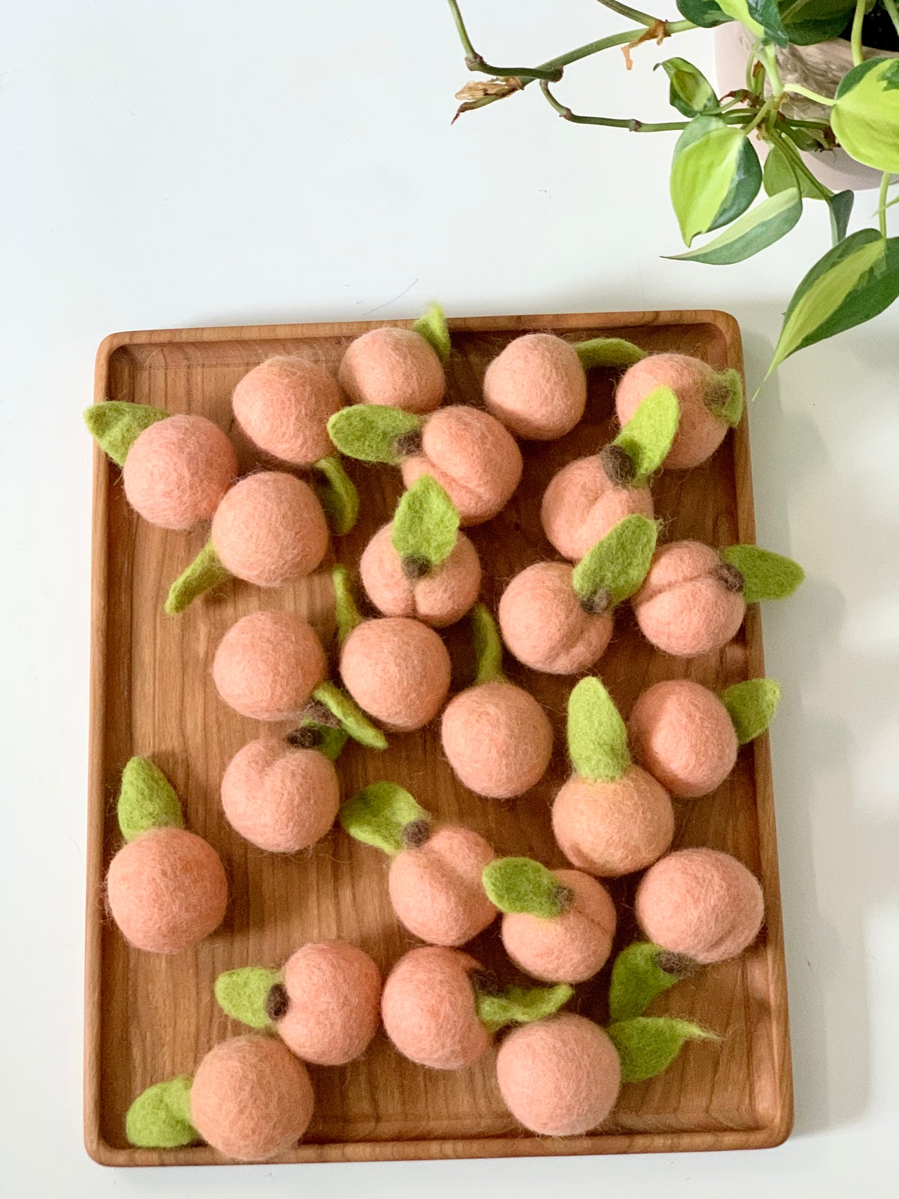 Wooden tray filled with small handmade felt peaches with green leaves on white table next to potted plant