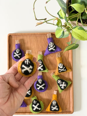 Handmade felt potion bottles in purple, orange, and green with skull and crossbones on wooden tray next to green plant