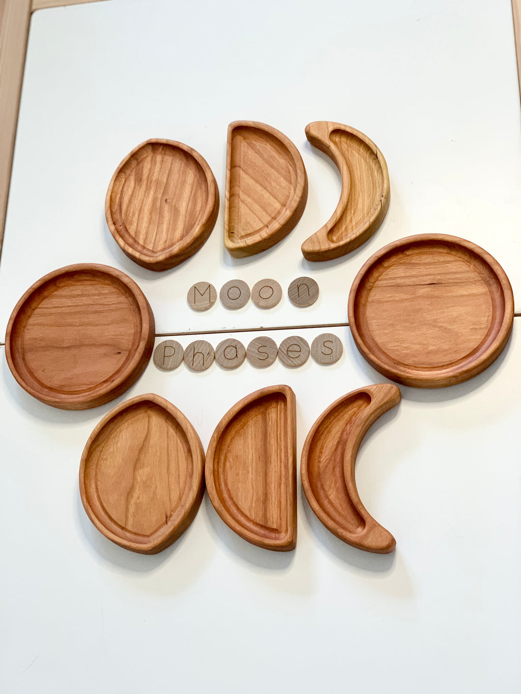 Set of wooden moon phase tinker trays arranged in circle with round wooden letter tiles spelling moon phases on white surface