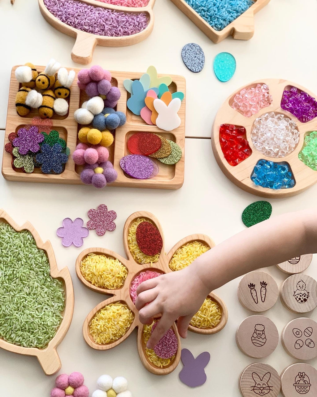Child’s hand arranging colorful textured craft materials in wooden trays shaped like flowers and leaves on a white surface