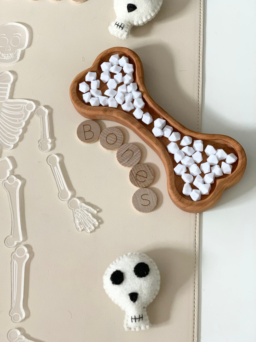 Bone-shaped wooden sensory tray filled with white foam cubes, wooden letter discs spelling bones, and felt skull toys on beige mat