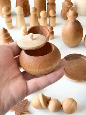 Hand holding open small round wooden container with various unfinished wooden shapes and figures in the background
