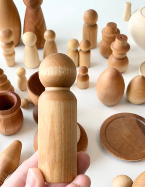 Hand holding smooth light natural wooden peg figure with various handcrafted wooden shapes and vessels in background