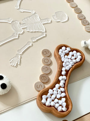 Wooden bone-shaped sensory tray filled with white star beads next to clear skeleton figure and wooden letter tiles spelling bones